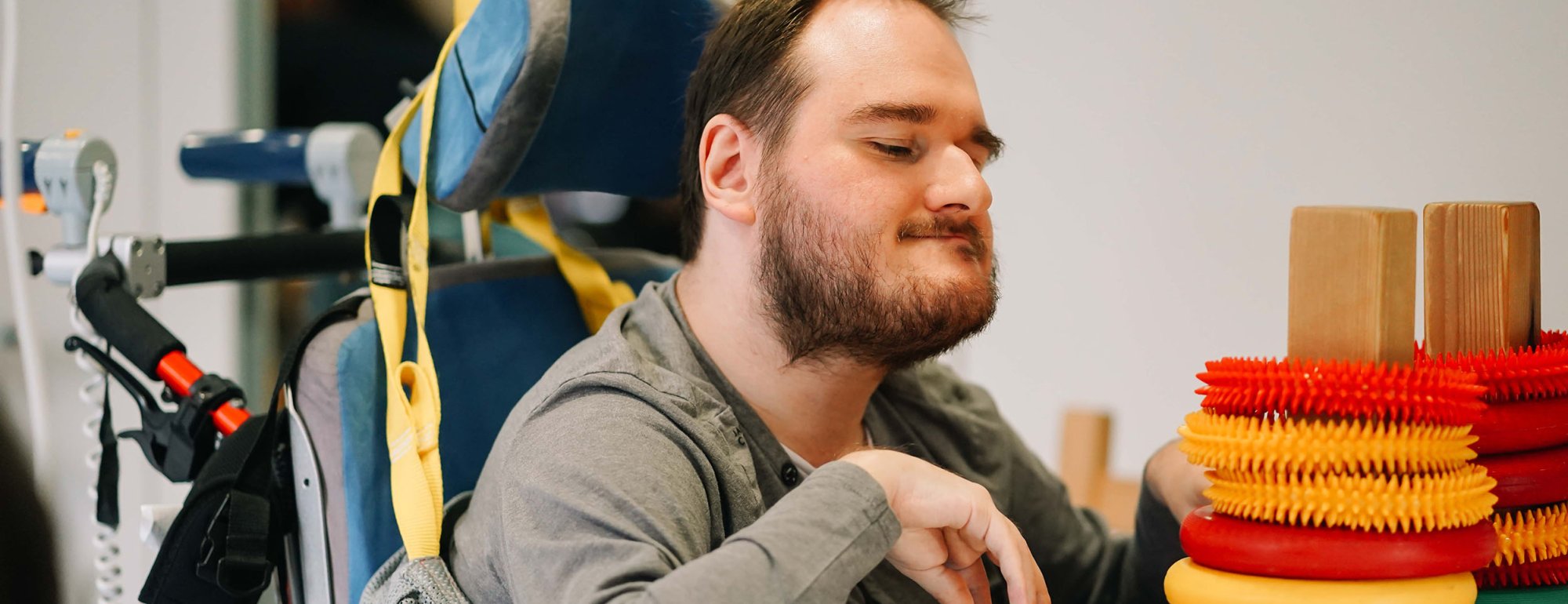 Ein junger Mann im Rollstuhl greift nach bunten Ringen, die über Holzstäbe auf dem Tisch vor ihm gereiht sind.