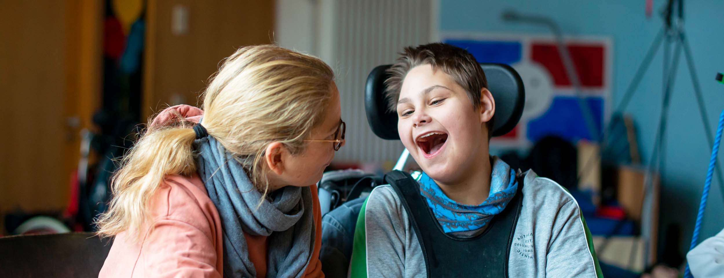 Im Vordergund ist rechts ein Junge im Rollstuhl zu sehen, der lacht. Links neben ihm sitzt eine Frau und hat sich zu ihm gedreht. Im Hintergrund ist ein Schrank, ein Heizkörper und eine blaue Wand zu sehen.
