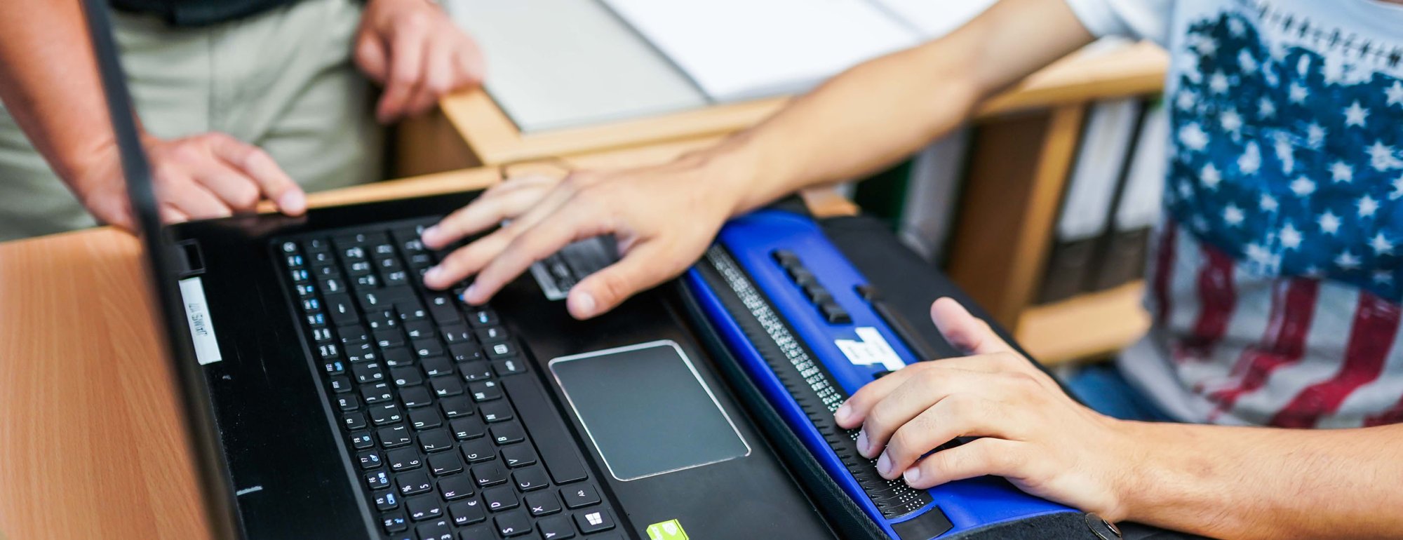 Blick von schräg oben auf einen Laptop, der auf einen Tisch steht. Vor dem Laptop steht eine Braille-Zeile, die den Text in Punktschrift ausgibt. Rechts angeschnitten ist ein Jugendlicher, der eine Hand auf der Laptop-Tastur hat und eine Hand auf der Braille-Zeile. Im Hintergrund ist ein erwachsener Mann von den Oberschenkeln bis zum Bauch zu sehen.