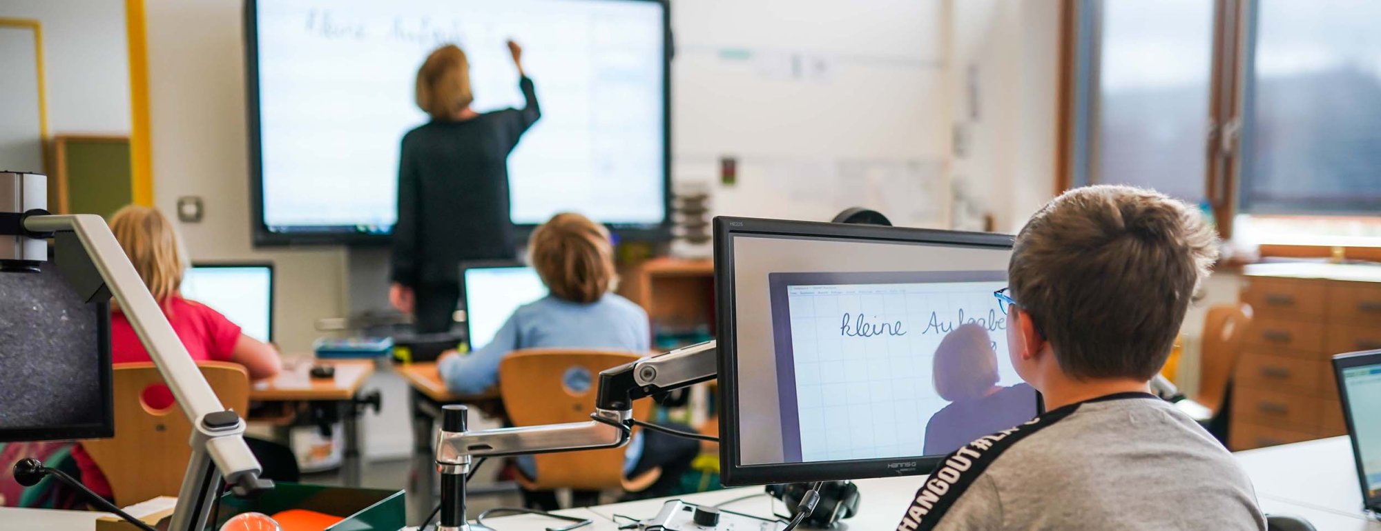 In einem Klassenzimmer sitzen mehrere Schüler vor einem Monitor, der sich vor ihrem Schultisch befindet. Eine Lehrerin schreibt auf eine digitale Tafel. Auf dem Monitor im Vordergrund ist die Lehrerin vor der digitalen Tafel vergrößert zu sehen.