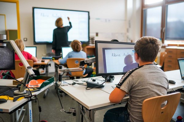 In einem Klassenzimmer sitzen mehrere Schüler vor einem Monitor, der sich vor ihrem Schultisch befindet. Eine Lehrerin schreibt auf eine digitale Tafel. Auf dem Monitor im Vordergrund ist die Lehrerin vor der digitalen Tafel vergrößert zu sehen.