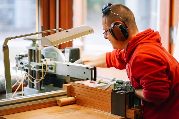 Ein junger Mann mit Brille und Gehörschutz sitzt an einer Werkbank am Fenster und arbeitet mit einer Maschine.