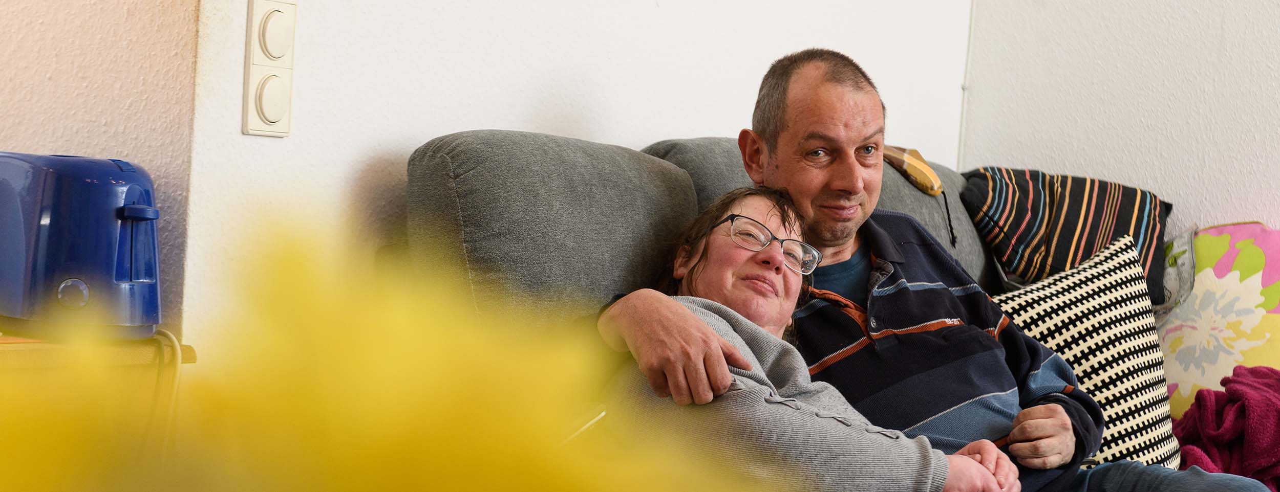 Ein Mann und eine Frau sitzen auf einem Sofa. Die Frau trägt eine Brille und hat sich an die Schulter des Mannes gelehnt. Beide schauen in Richtung des Betrachters und lächeln.