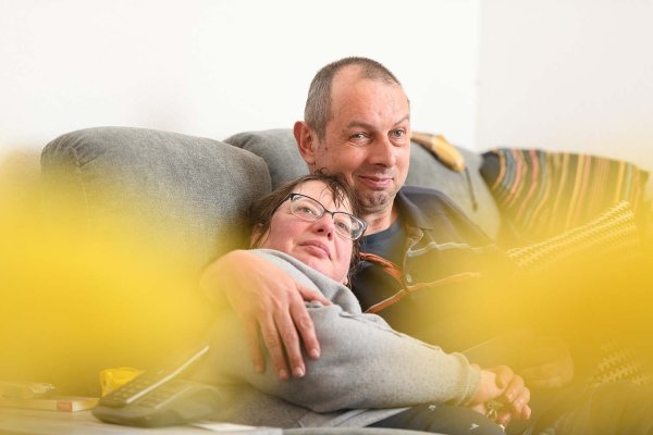 Ein Mann und eine Frau sitzen auf einem Sofa. Die Frau trägt eine Brille und hat sich an die Schulter des Mannes gelehnt. Beide schauen in Richtung des Betrachters und lächeln.