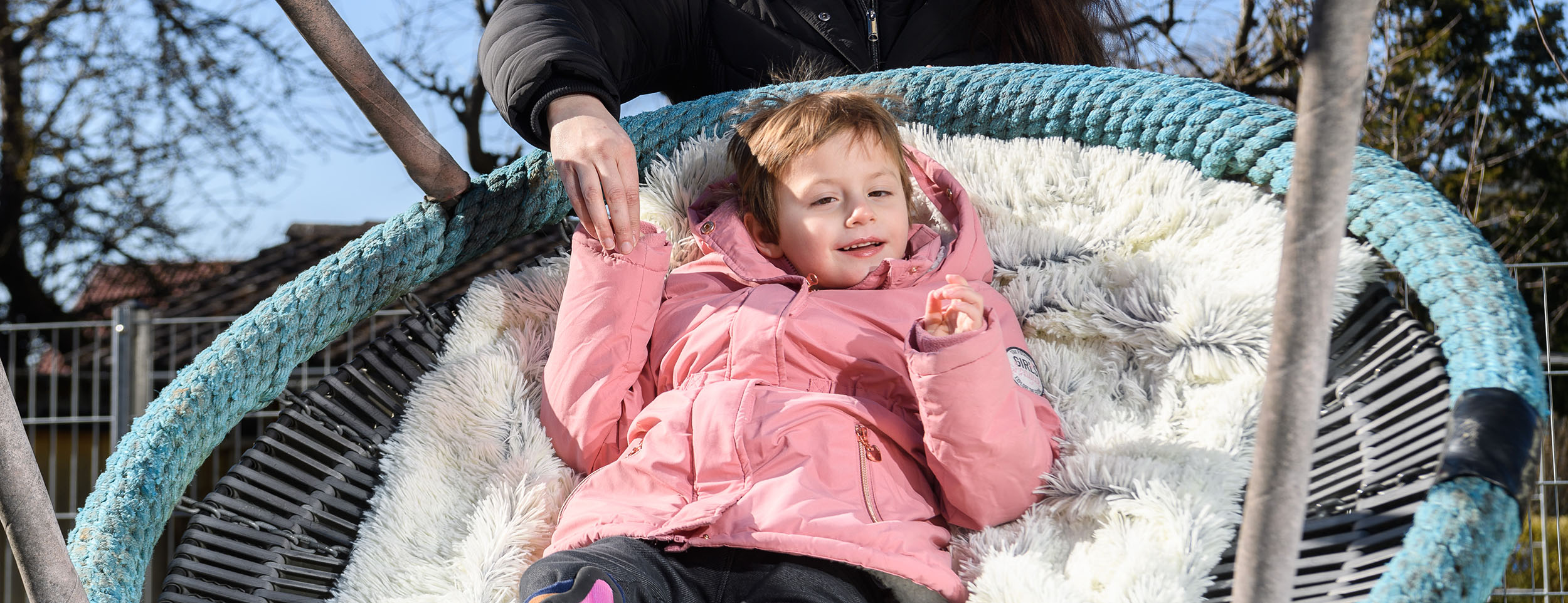 Auf einer Vogelnest-Schaukel liegt ein kleines Mädchen in einer rosafarbenen Jacke auf einer kuscheligen weißen Decke. Hinter der Schaukel steht eine junge Frau mit schwarzer Jacke, die mit einer Hand die Hand des Mädchen hält. Beide lachen. Im Hintergrund ist ein wolkenfreier blauer Himmel zu sehen.