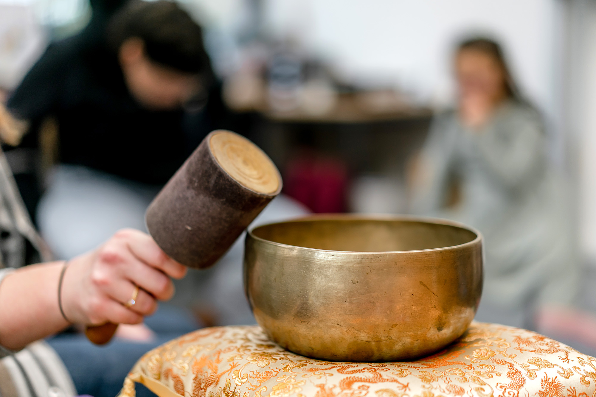 Im Bild ist eine Klangschale auf einem Kissen abgebildet, links davon ein Klöppel, der gerade die Schale anschlagen will.