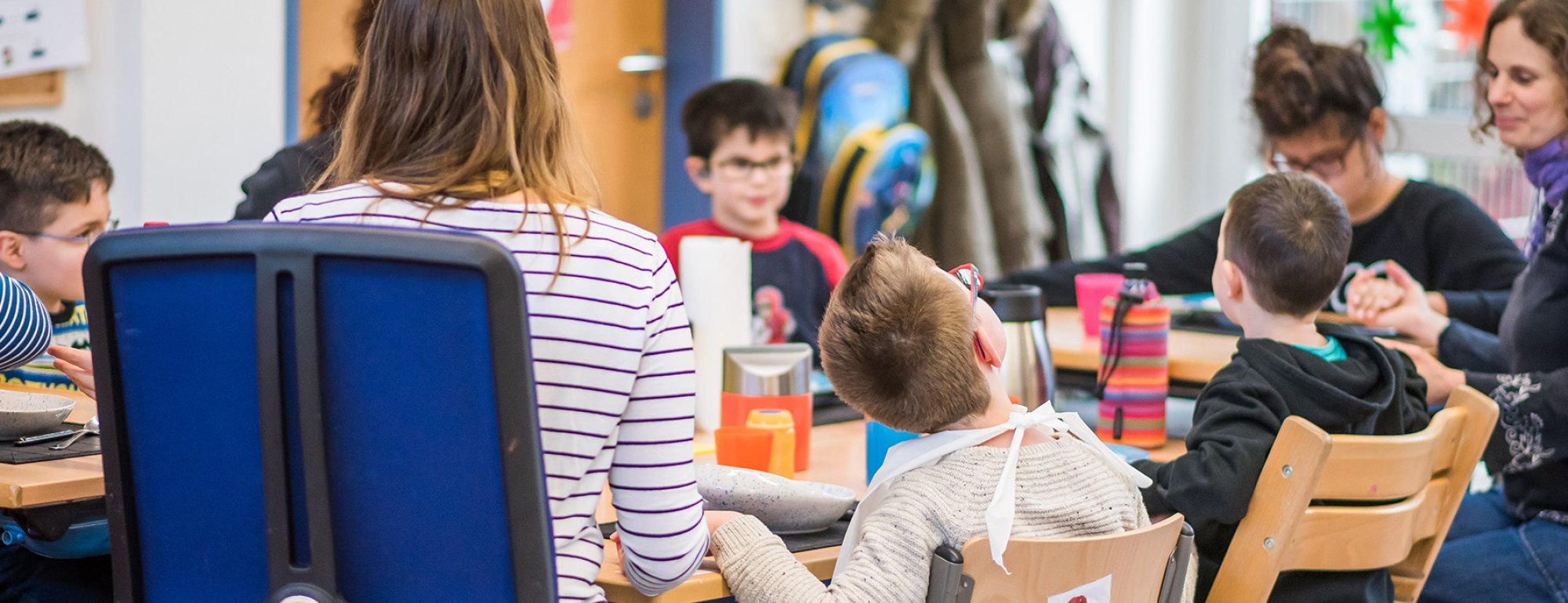 Eine Gruppe von Kindern und Jugendlichen sitzt mit zwei Erzieherinnen um einen Mittagstisch. Alle haben sich die Hände gereicht und wünschen sich einen guten Appetit.