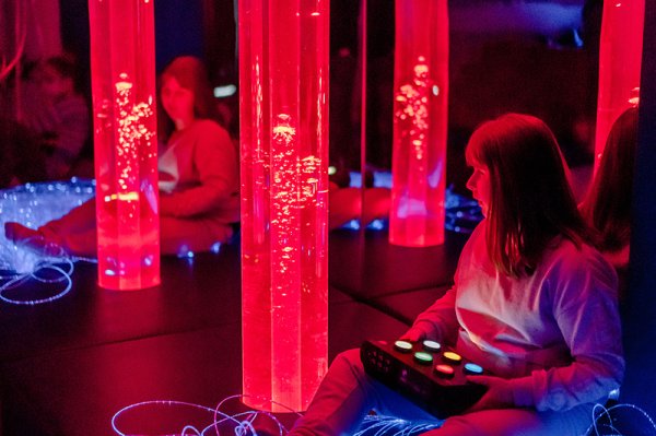 Ein junges Mädchen sitzt zwischen Plexiglasröhren und betrachtet die mit Wasser gefüllten, rot beleuchteten Röhren, in denen Luftblasen aufsteigen.