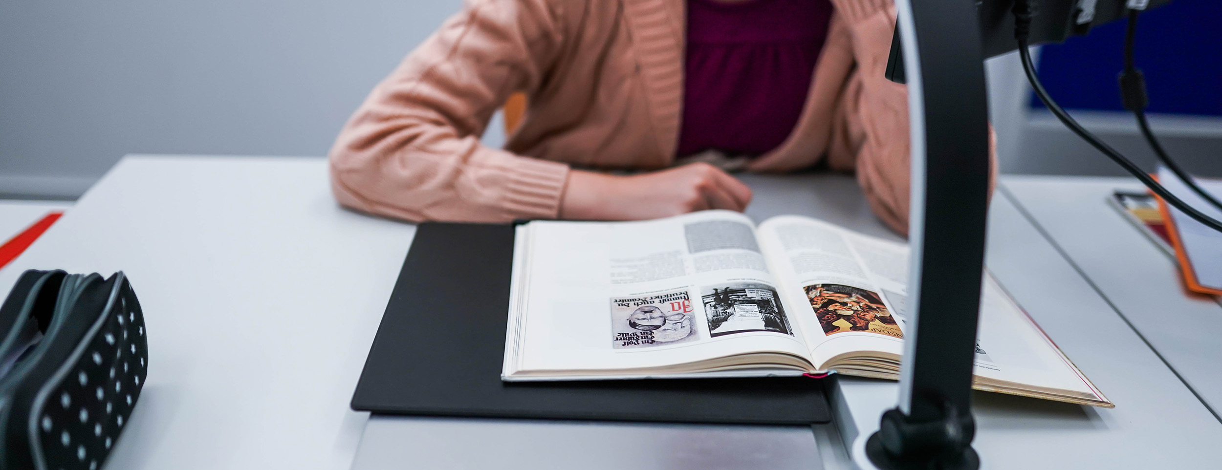 Eine Schülerin hat vor sich auf dem Tisch ein offenes Buch liegen, das sie mithilfe eines Bildschirmlesegeräts liest.