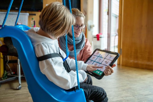 Eine Mitarbeitende des Blindeninstituts Rückersdorf hilft einem Jungen in einer Schaukel bei Lernübungen mit einer digitalen Tafel.