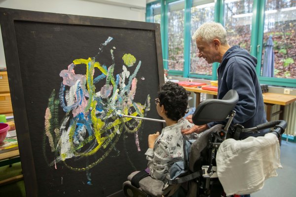 Ein im Rollstuhl sitzendes Mädchen malt auf eine Leinwand. Unterstützt wird sie dabei vom Leiter des Kunsch-D-Projekts.