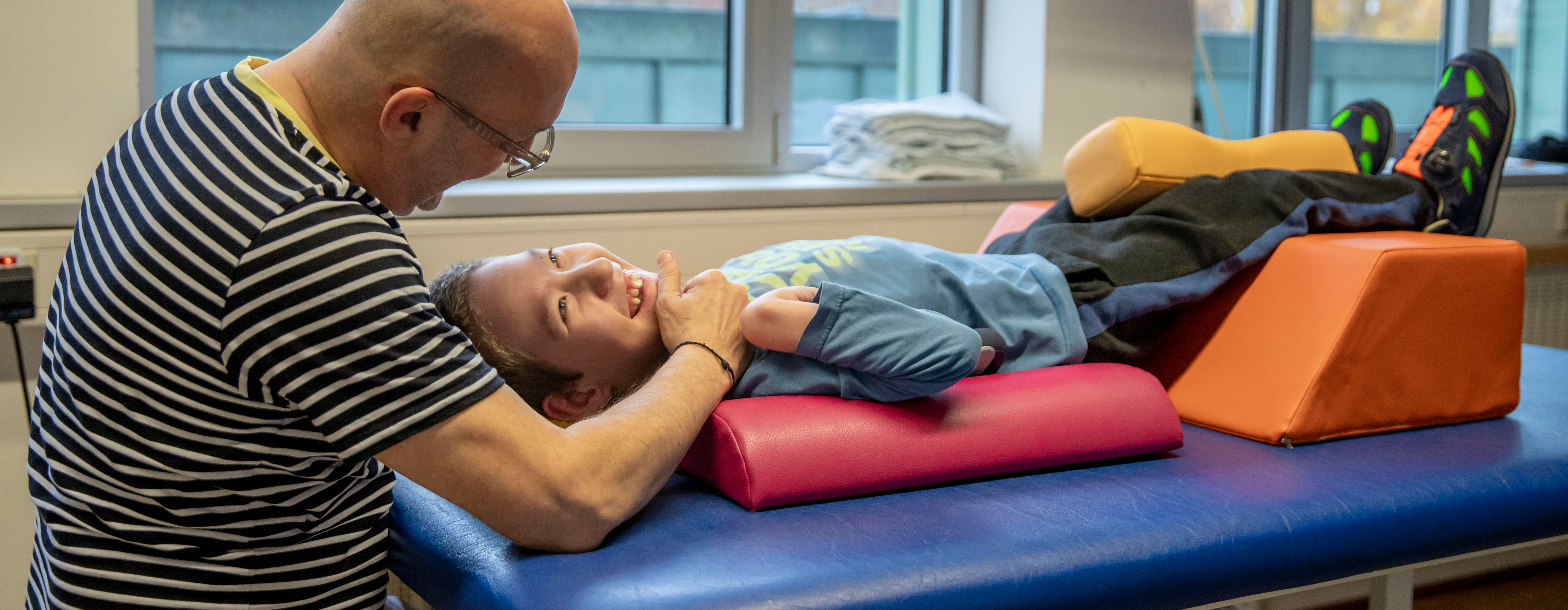 Ein Mitarbeiter der Medizinischen Therapie behandelt einen lachenden Jungen auf einer Liege.