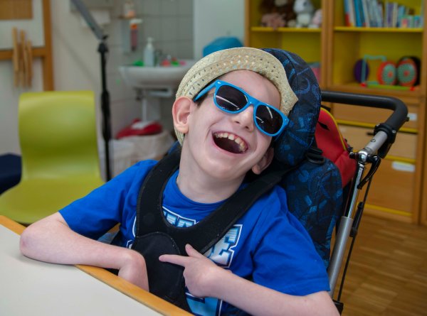 Das Bild zeigt einen lachenden Jungen mit Sonnenbrille im Rollstuhl sitzend.