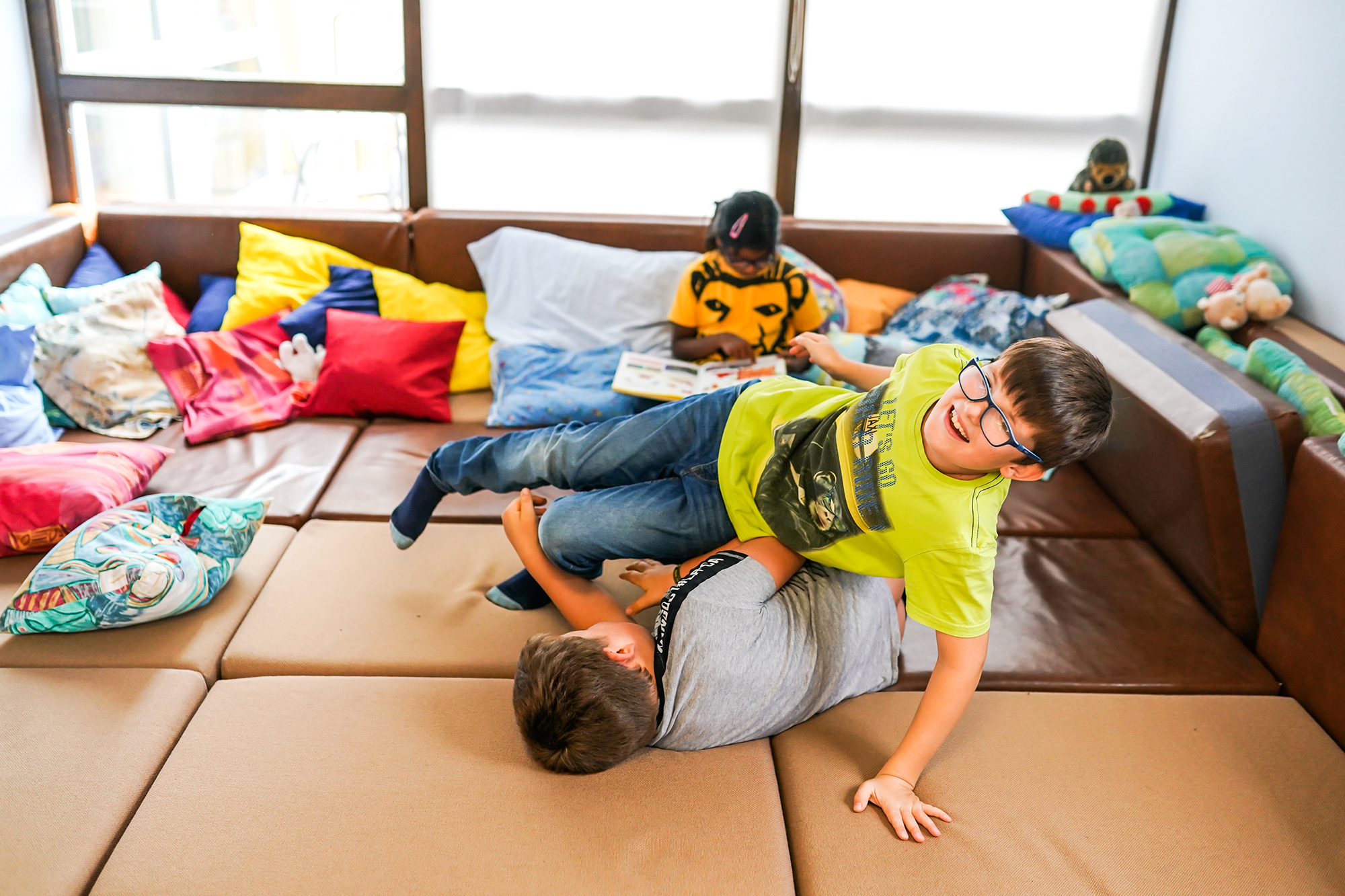 Zwei Jungen raufen auf einer Schaumstoffmatte, im Hintergrund sitzt ein Mädchen und liest.