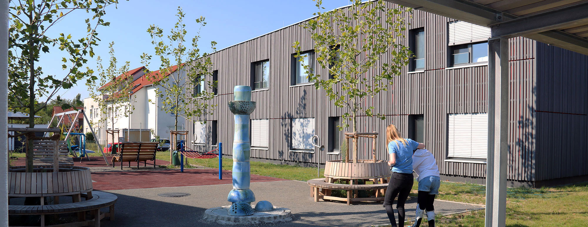 Ein überdachter offener Gang führt durch ein Gartengelände vom Wohnhaus in die Förderstätte. Im Gang sind ein Rollstuhlfahrer und zwei weitere Menschen zu sehen. 
