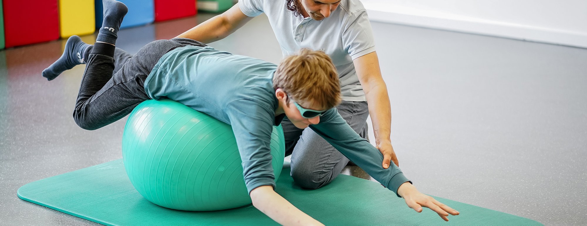 Ein Physiotherapeut übt mit einem Jungen, der auf einem Therapieball liegt, das Gleichgewicht zu halten.