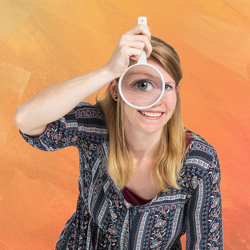 Eine junge Frau hält eine Lupe verkehrt herum vor ihr Auge. Das Auge wird durch das Lupenglas hindurch stark vergrößert.