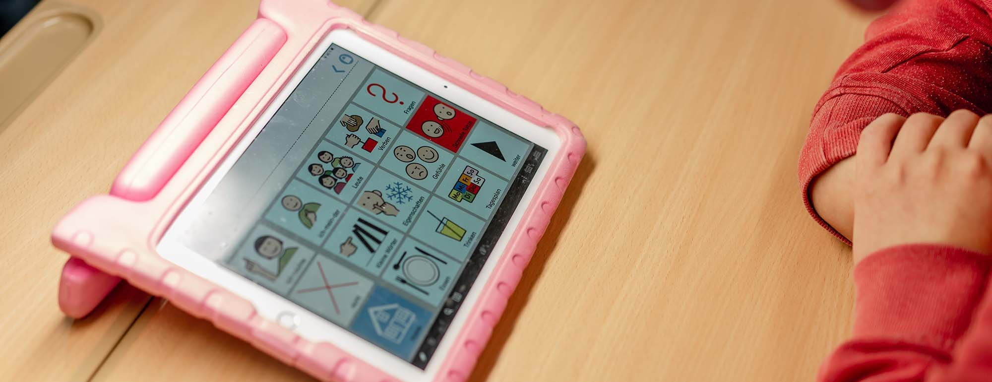 Auf einem Schultisch steht ein rosafarbiges Tablet, auf das eine Schülerin blickt. Auf dem Bildschirm sind verschiedene Symbole und Grafiken zu sehen.