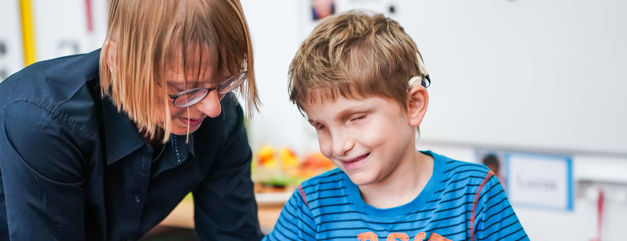 Eine erwachsene Frau mit Brille und kinnlangen Haaren beugt sich freundlich zu einem Jungen. Der Junge hat ein Cochlea-Implantat am Ohr und scheint sehbehindert oder blind zu sein. Er lächelt leicht und trägt ein blaues T-Shirt mit schwarzen Streifen. Beide befinden sich in einem freundlichen Raum, vermutlich einem Klassenzimmer oder Förderraum. Die Frau unterstützt den Jungen bei einer Aufgabe. Die Szene wirkt ruhig, wertschätzend und aufmerksam