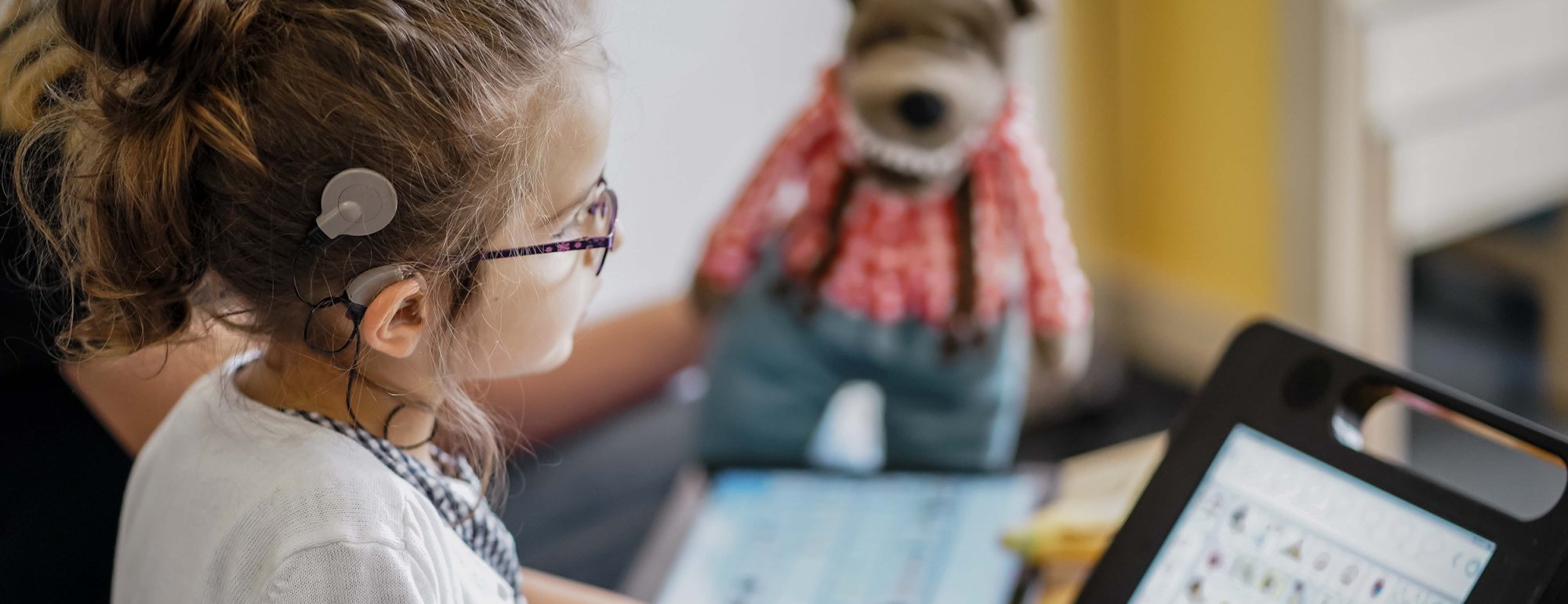 Ein Kind mit einem Cochlea-Implantat sitzt an einem Tisch und schaut auf ein Tablet, auf dem Symbole und Bilder zu sehen sind. Vermutlich handelt es sich um eine unterstützende Kommunikations-App. Im Hintergrund ist eine erwachsene Person zu erkennen, die auf das Tablet zeigt. Auf dem Tisch, auf dem das Tablet steht, sitzt außerdem ein Stofftier. Die Szene wirkt wie ein inklusives Lern- oder Therapieumfeld, in dem das Kind mithilfe digitaler Hilfsmittel kommuniziert.