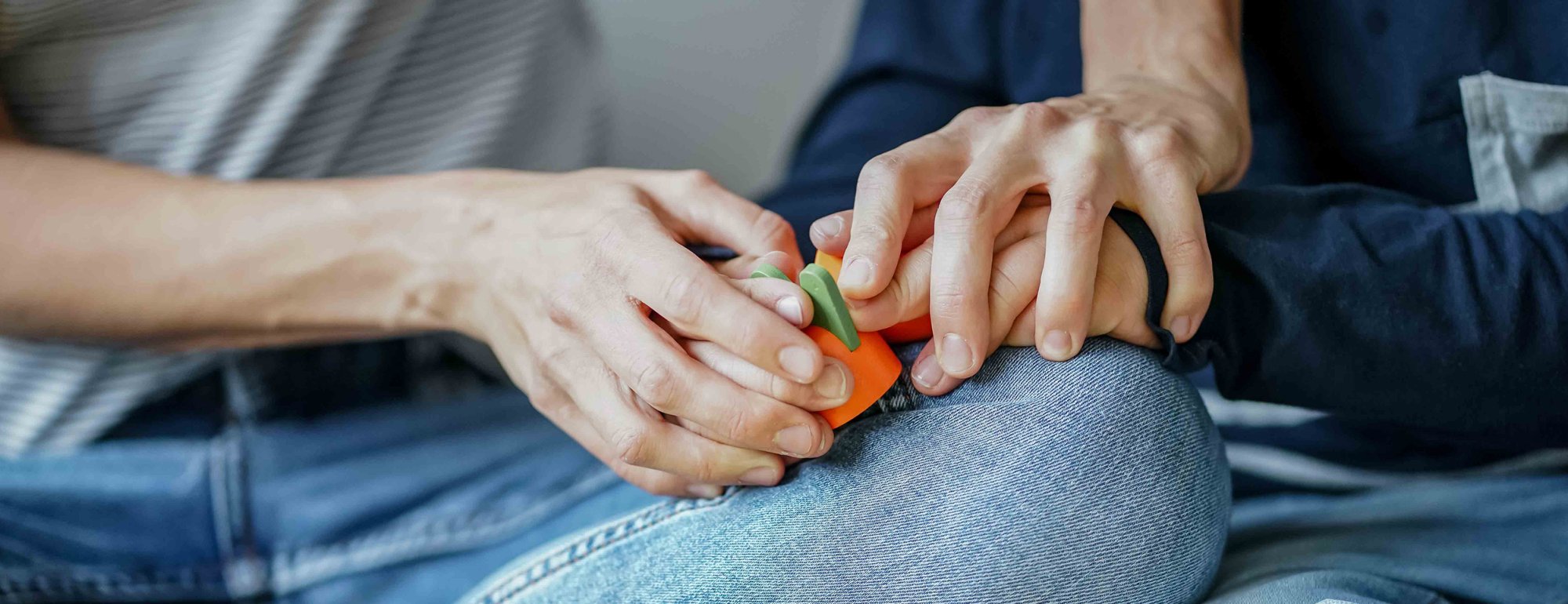 Hände zweier Personen berühren sich sanft und halten gemeinsam ein buntes Spielzeug, während die Hände auf dem Oberschenkel einer Person ruhen. Das Bild vermittelt Nähe, Vertrauen und unterstützende Begleitung in einer ruhigen, geschützten Atmosphäre.