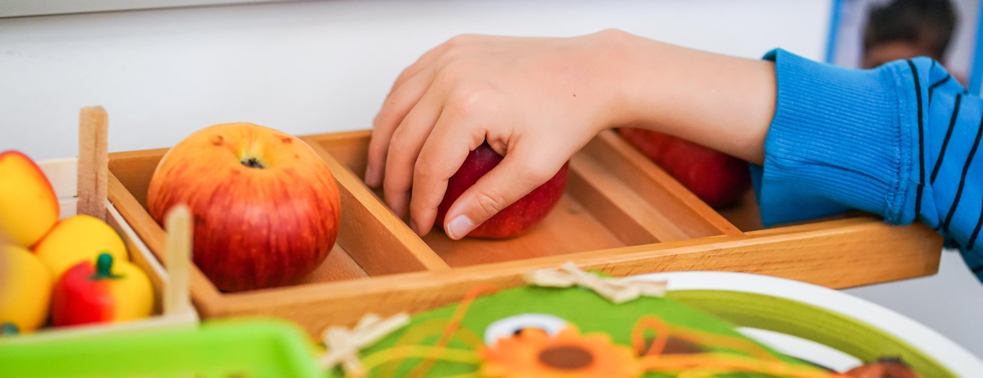 Ein Kind greift mit der Hand nach einem roten Apfel, der in einem Holzkästchen zwischen anderen Früchten liegt.