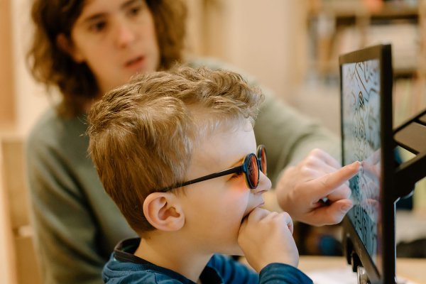 Ein Junge sieht mit wenig Abstand auf einen Monitor, der schwarze Linien eines Ausmalbildes zeigt. Er trägt ein Brille mit rötlichen Gläsern und hat die Hand im Mund. Im Hintergrund ist eine Frau, die mit ihrem Zeigefinger auf den Monitor deutet.