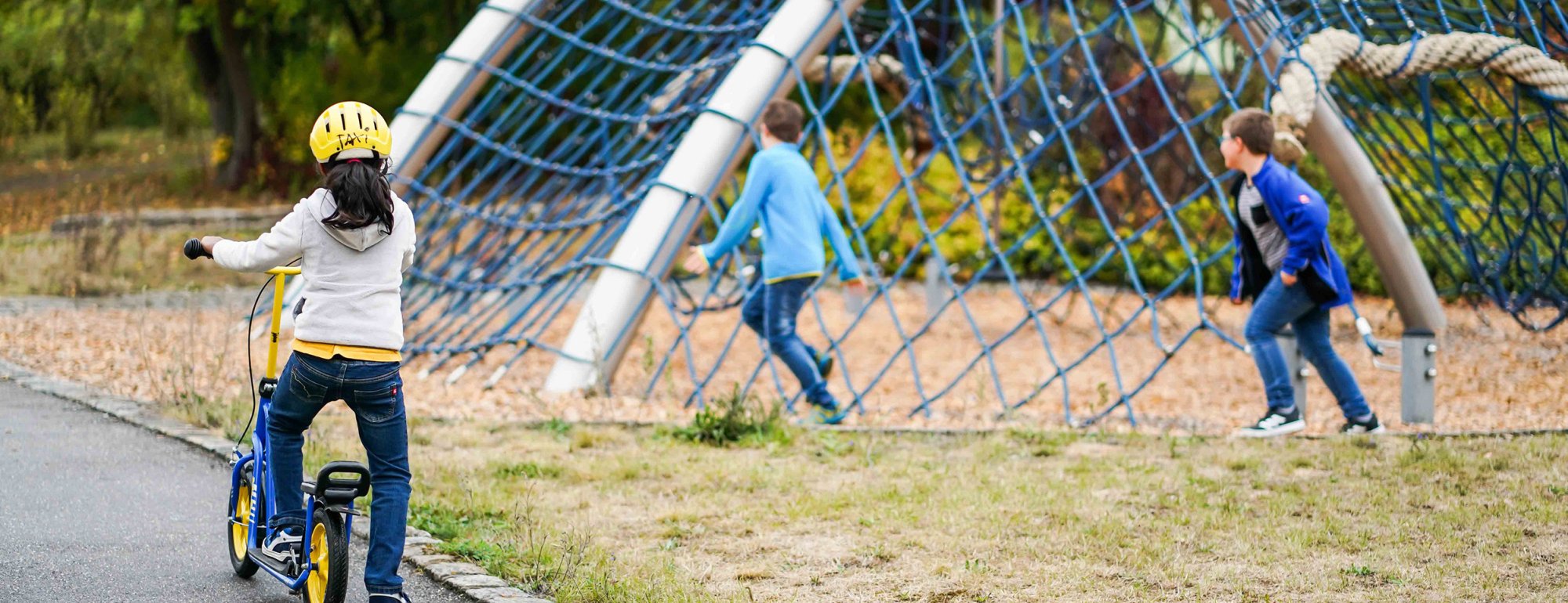 Ein Mädchen auf einem Roller sieht zu zwei Jungen an einem großen Kletterspielgerät hinüber.